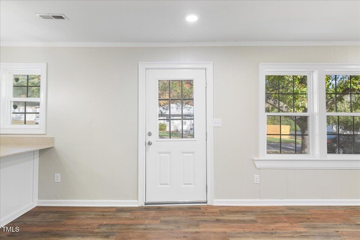 A room with a white door and three windows. The walls are light-colored, and there is a wooden floor. Sunlight is streaming in through the windows, and a counter is partially visible on the left side.