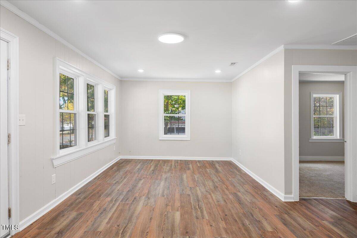 An empty room with light beige walls and wood flooring, featuring large windows allowing natural light. A doorway on the right leads to another room with carpeted flooring. Ceiling lights are present, and the space feels bright and inviting.