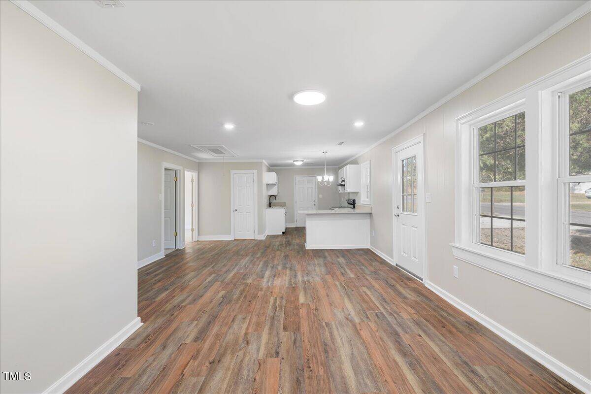Interior of a modern, empty room with wood-style flooring. The room has light-colored walls, a white door, and two large windows on the right. The view extends to an open kitchen with white cabinets and a central light fixture on the ceiling.