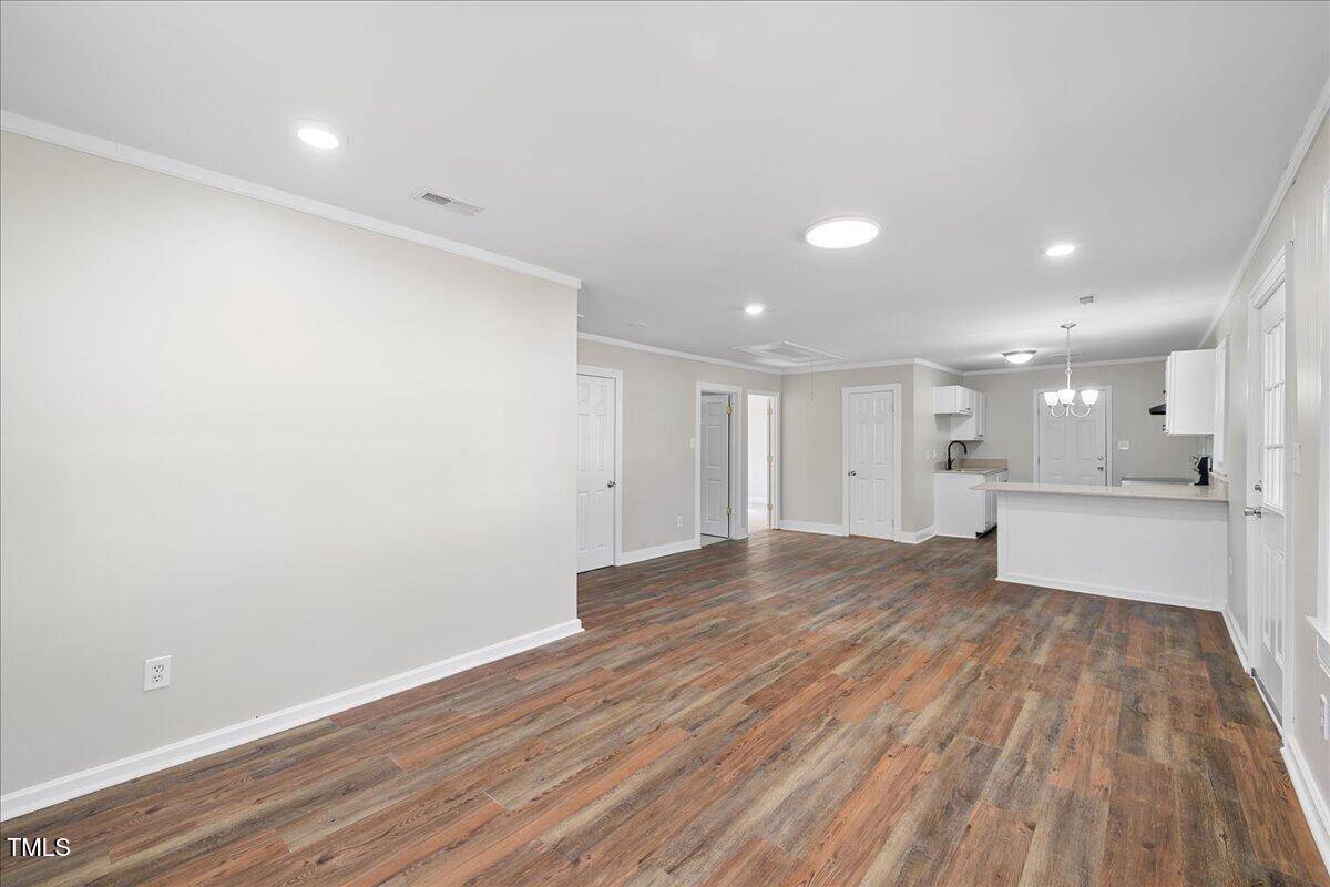 Spacious open-plan living area with wood flooring, white walls, and recessed lighting. The room leads to a kitchen with white cabinets and an island. Several doors are visible, suggesting multiple rooms and storage spaces.
