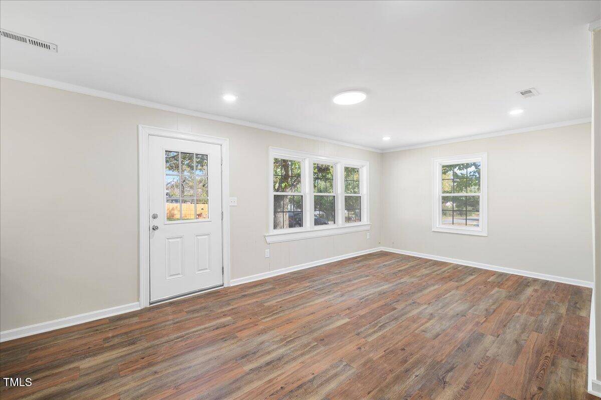 A bright, empty room with light-colored walls and wood flooring. The space has a white door and three large windows along one wall, letting in natural light. Recessed ceiling lights are turned on.