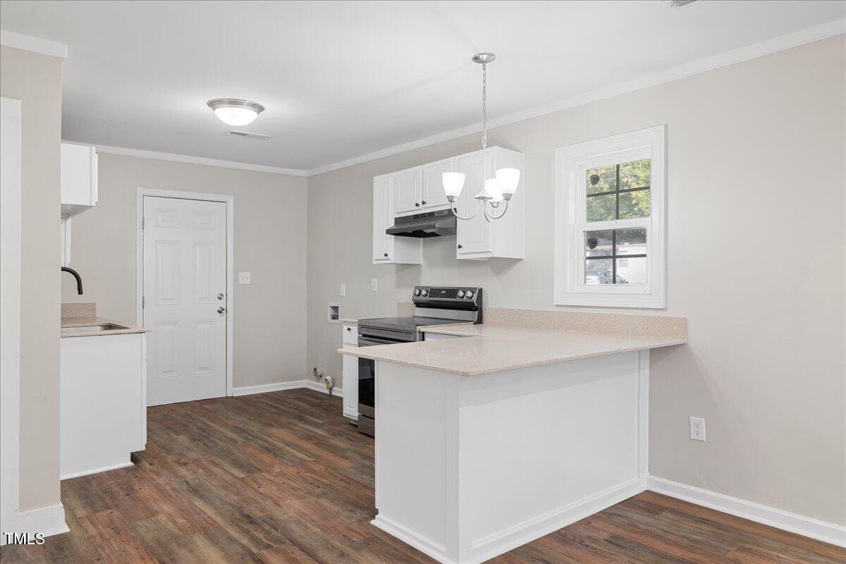 A modern kitchen with white cabinets, a black stove, and a beige countertop. A small kitchen island is present beneath a hanging light fixture. The room features wood-look flooring and a window letting in natural light.