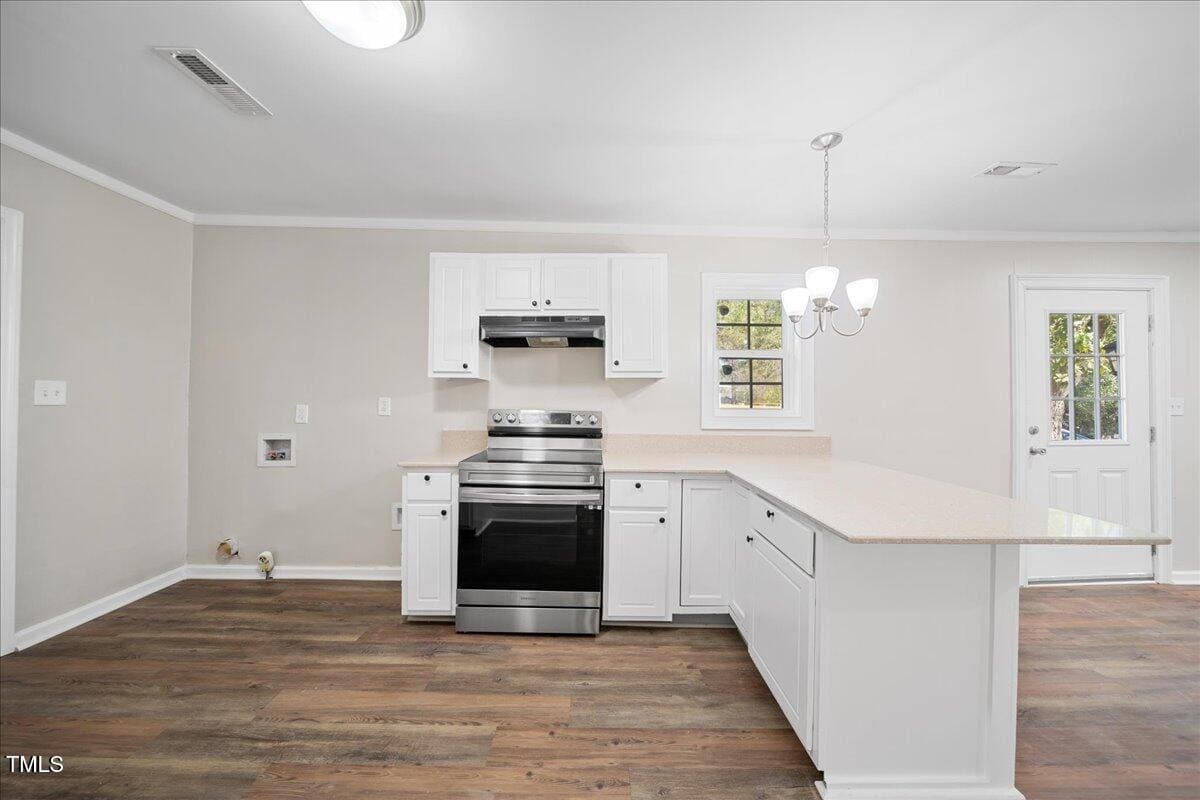 A modern kitchen with white cabinets, stainless steel stove and range hood, and light-colored countertops. The room features a small chandelier, wooden flooring, and a door with a window leading outside.