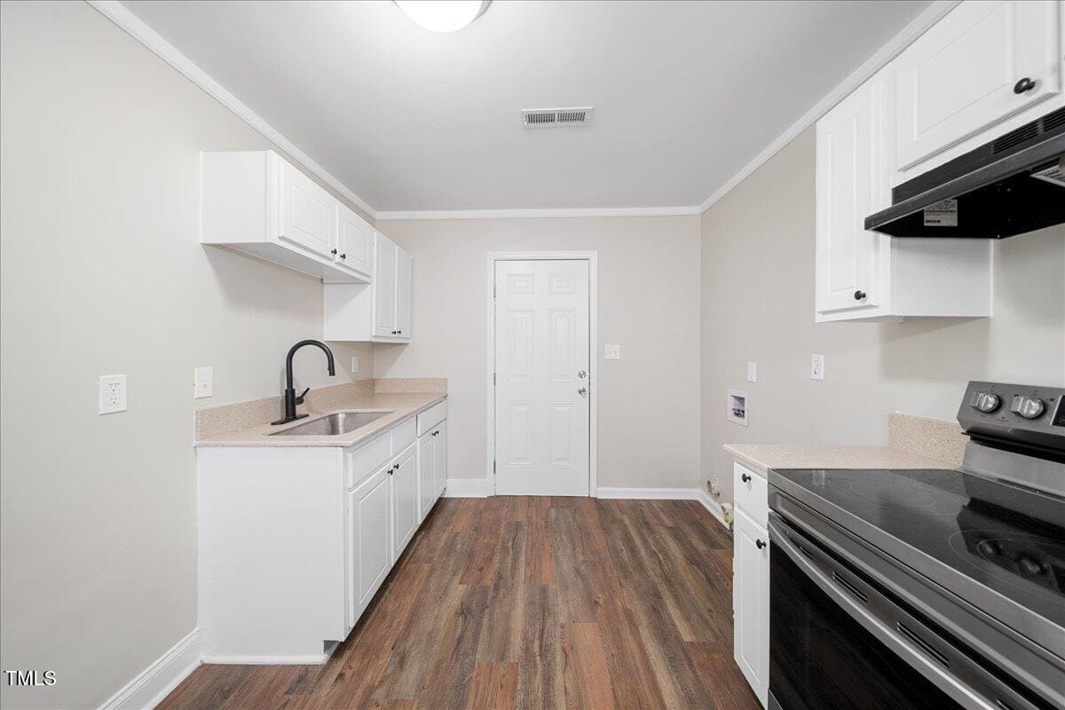 A modern kitchen with white cabinets, a black faucet, light countertops, and wood floor. A white door is in the background next to an electric stove with a black top. The walls are painted light gray.
