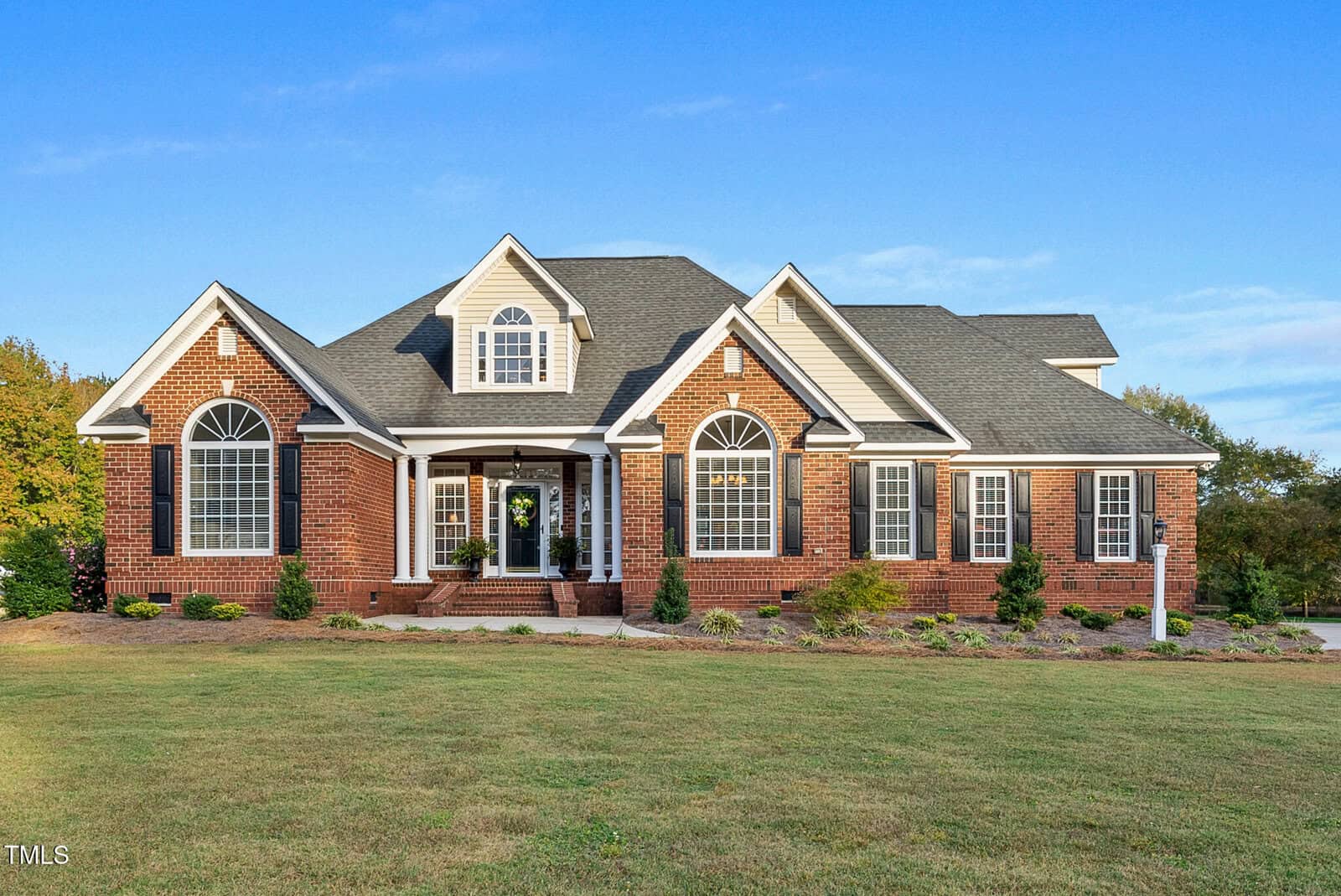 A large, two-story brick house with white trim, multiple arched windows, a gray shingled roof, and a manicured lawn, set against a blue sky and trees in the background.