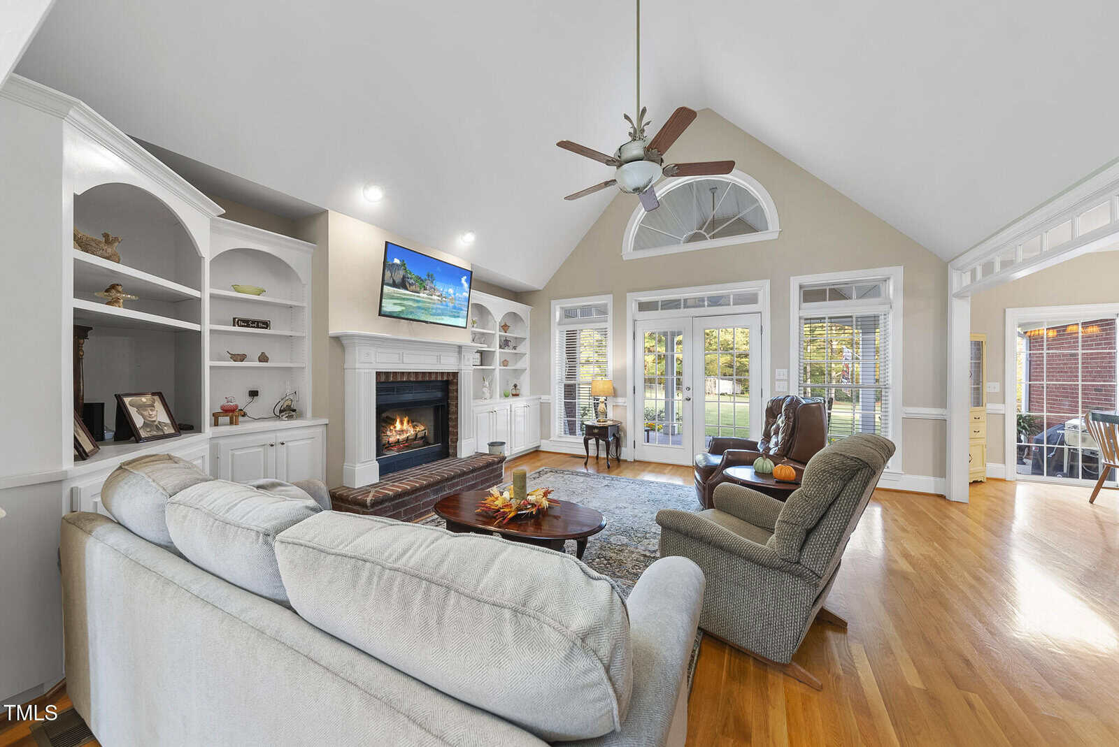 A spacious, bright living room with vaulted ceiling, ceiling fan, fireplace, built-in shelves, a TV above the mantel, large windows, and cozy seating, including a sofa and armchair on a hardwood floor.