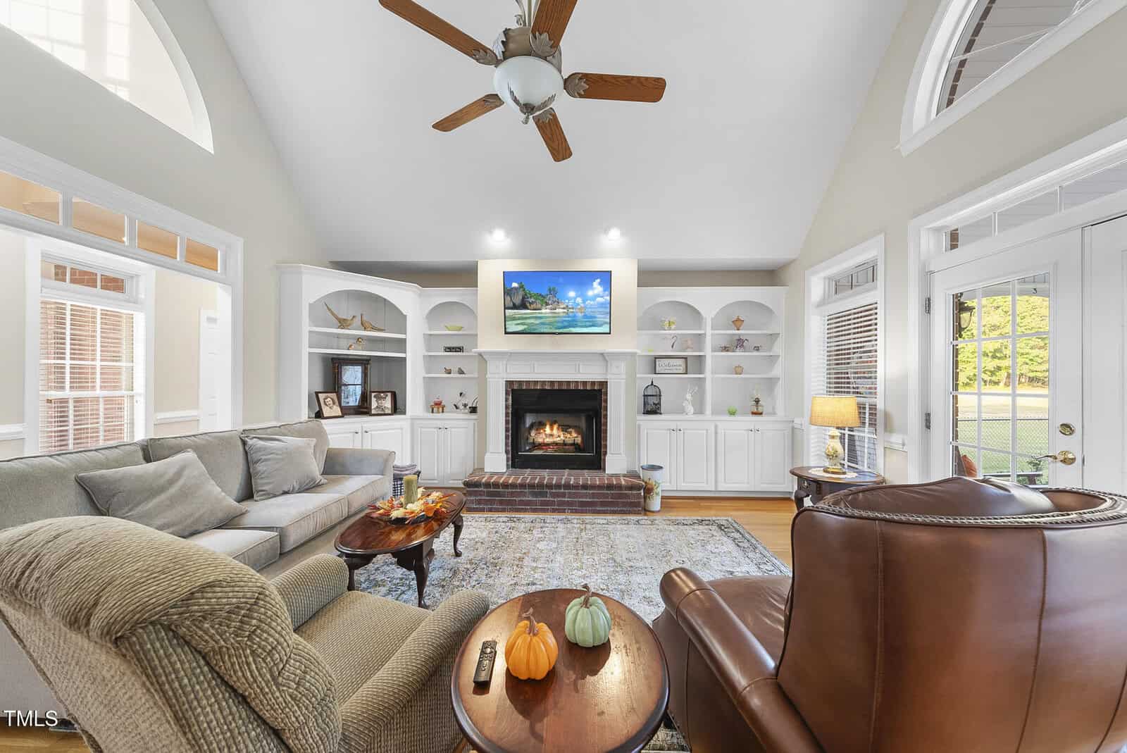 A bright living room with vaulted ceilings, a ceiling fan, cozy seating, a fireplace with a TV above it, built-in shelves, and large windows letting in natural light. A small table holds two decorative pumpkins.