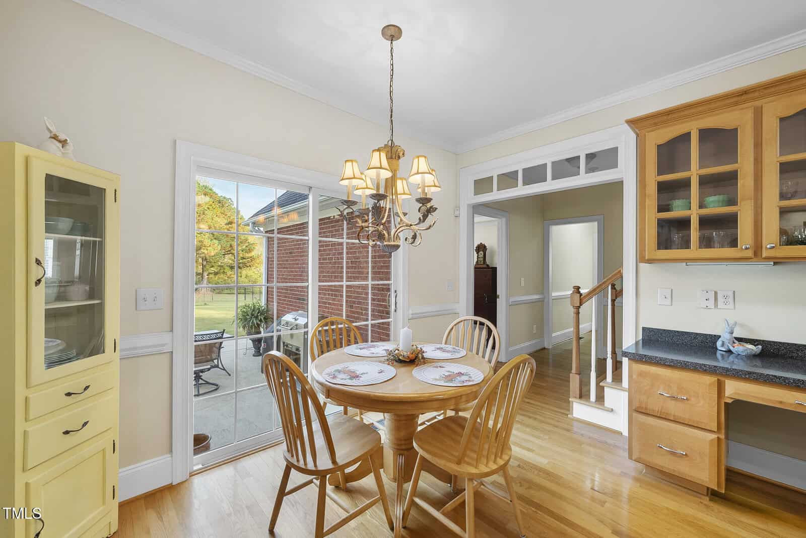 A cozy dining area with a wooden table and four chairs sits by large glass doors leading to a patio. The space features hardwood floors, a chandelier, a yellow cabinet, and wooden kitchen cabinets with glass doors.