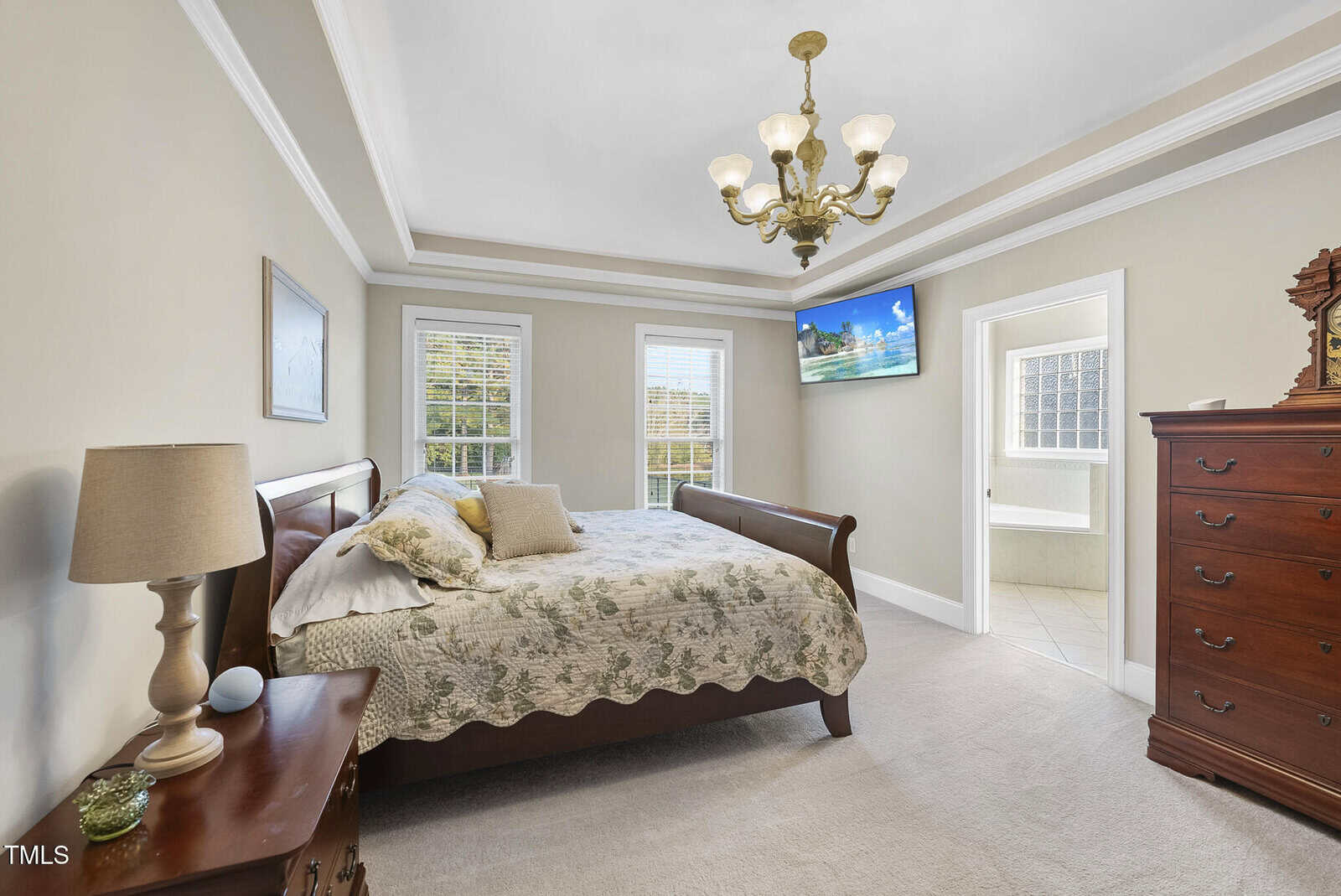 A bright bedroom with a wooden sleigh bed, floral bedding, two windows, a chandelier, a TV mounted on the wall, and a dresser. An open door leads to a bathroom with tiled floors.