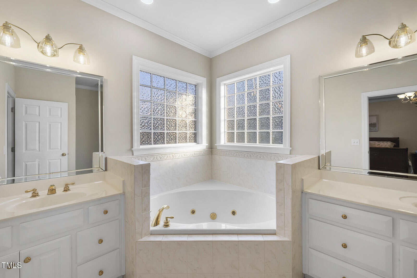 Spacious bathroom with a white corner bathtub, gold fixtures, two vanities with mirrors and lights, large textured glass windows, and white cabinetry. Walls are painted light beige.