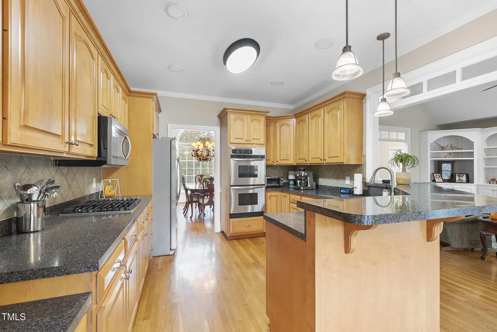 Spacious kitchen with light wood cabinets, dark granite countertops, stainless steel appliances, double ovens, and a large island; dining room with chandelier visible through an open doorway.