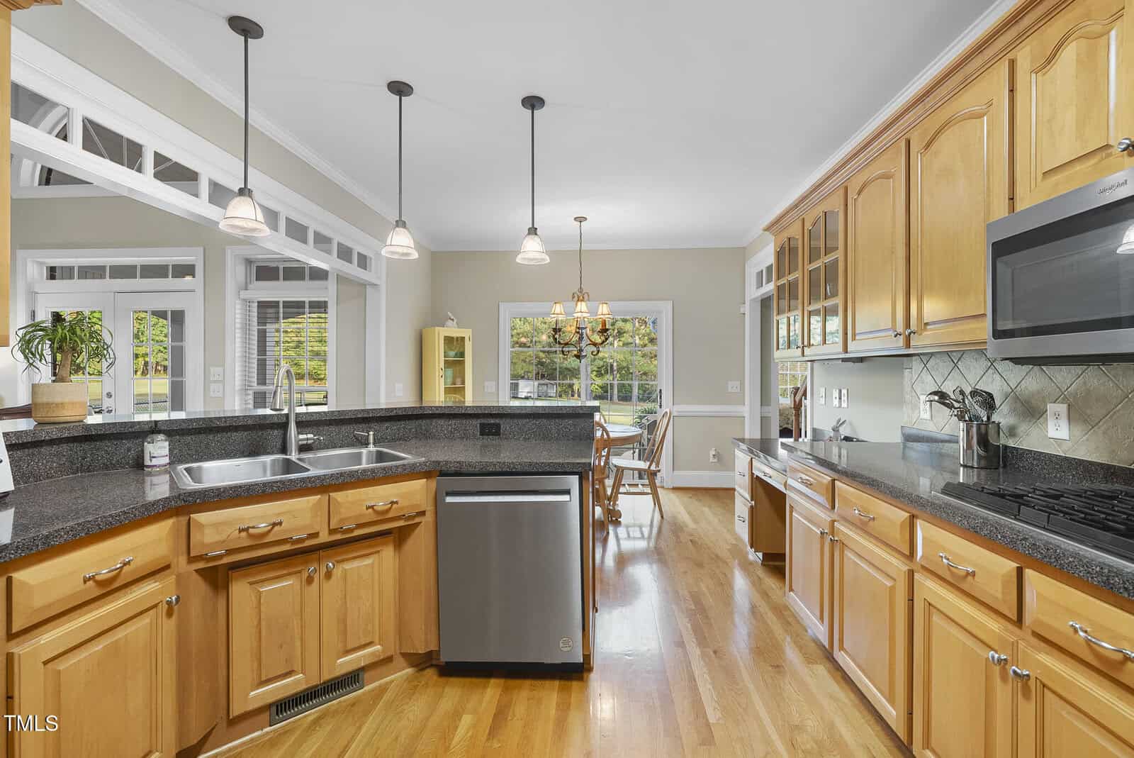 Spacious kitchen with wooden cabinets, black countertops, stainless steel appliances, and a breakfast bar. Large windows and hanging pendant lights create a bright, open atmosphere leading to a dining area.