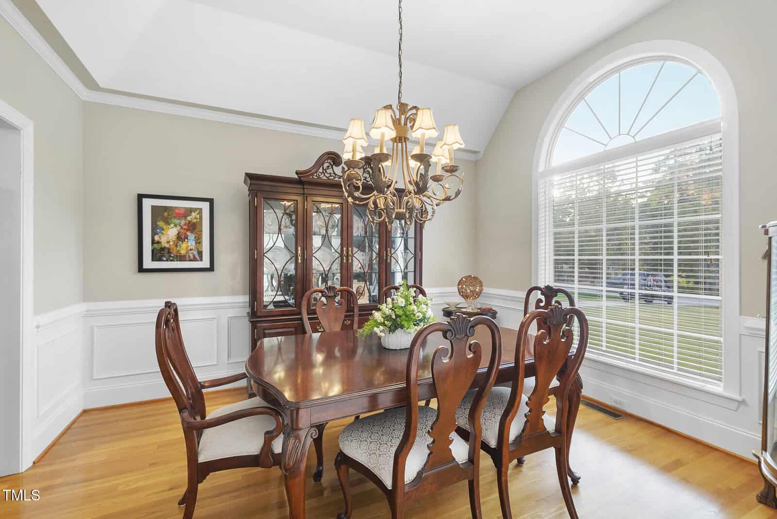 Elegant dining room with a wooden table and six matching chairs, a chandelier above, a glass-fronted china cabinet, wall art, and a large arched window letting in natural light. The walls are light and the floor is wood.