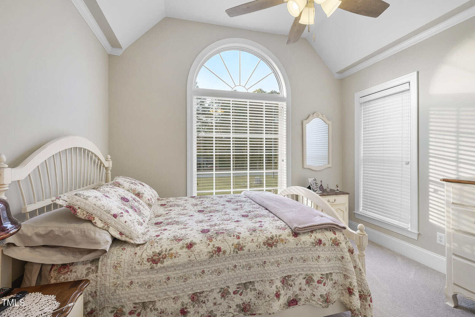Bright bedroom with vaulted ceiling, arched window, ceiling fan, floral bedding on a white bed, dresser with mirror, and lots of natural light from two large windows.