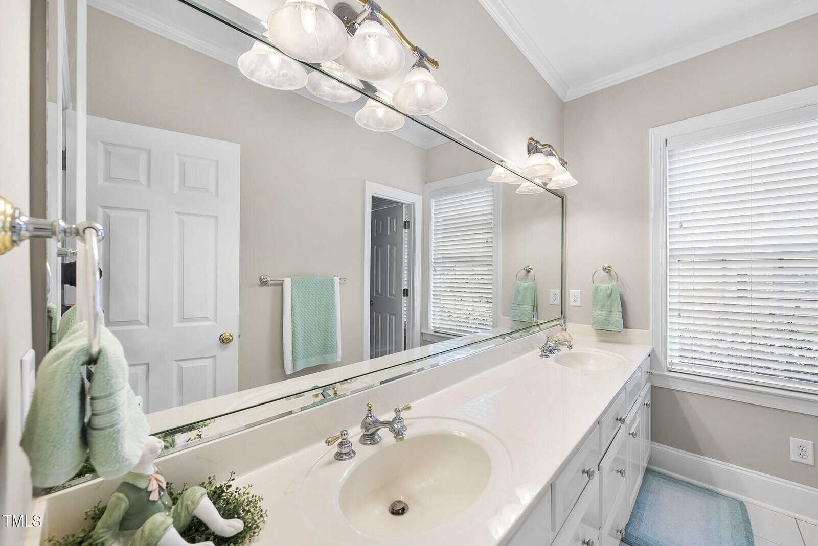Bright bathroom with a double sink vanity, large wall mirror, silver faucets, white cabinets, mint green towels, rabbit plush, and window with white blinds letting in natural light.