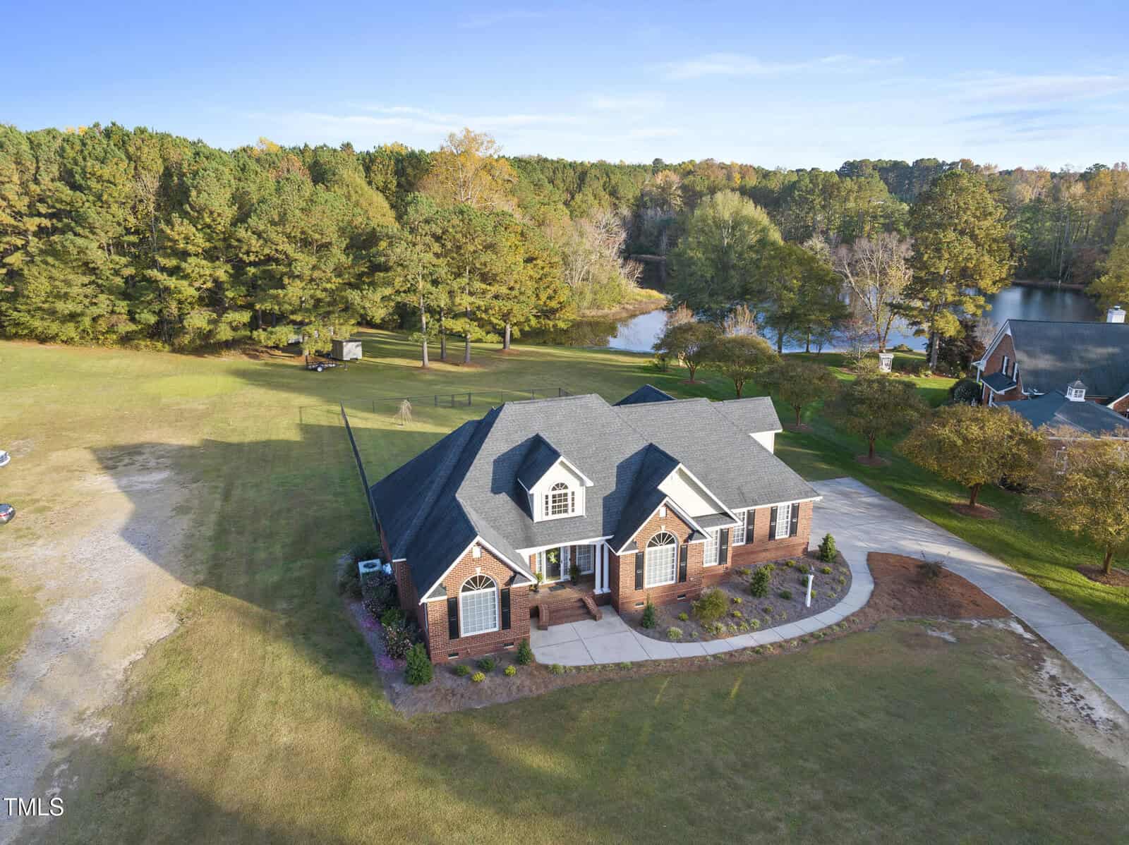 Aerial view of a large brick house with a dark roof, surrounded by a spacious grassy yard, trees, and a nearby lake, under a clear blue sky.