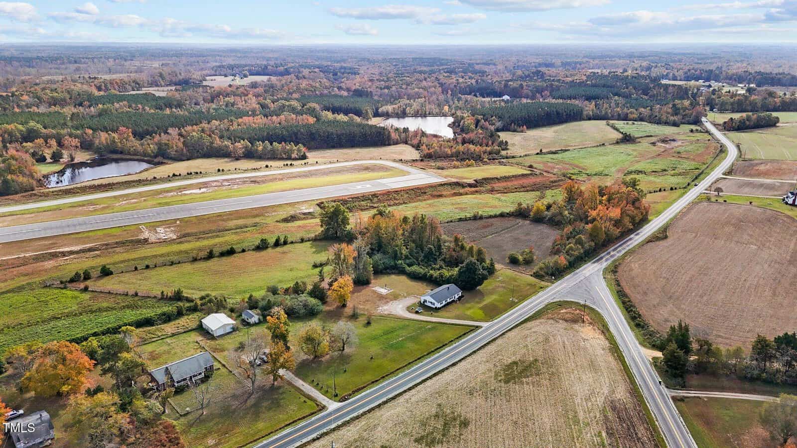 Aerial view of a rural landscape featuring an airstrip, fields, and a few houses. Roads intersect in the foreground. Trees, some with autumn foliage, and a distant lake can be seen under a partly cloudy sky.