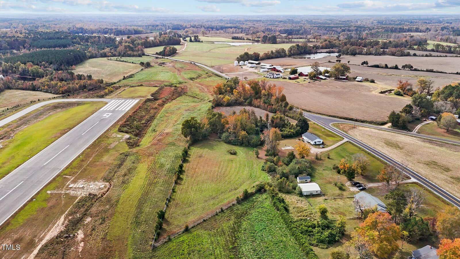 A rural landscape with an airstrip on the left, surrounded by fields and scattered trees. Buildings and a few roads are visible in the distance under a cloudy sky. Autumn foliage colors the scene.