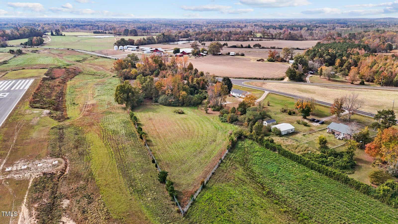 Aerial view of a rural area with fields and scattered trees. A triangular fenced pasture is in the foreground, and several homes and barns are visible. A small runway is on the left, and the landscape stretches into the horizon under a clear sky.