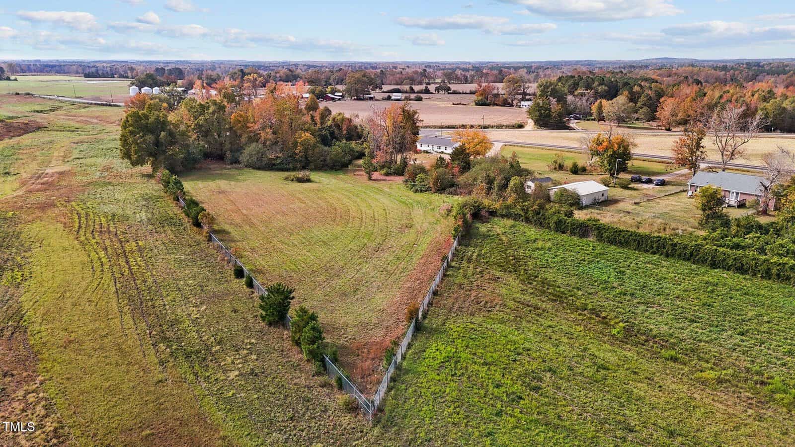 Aerial view of a triangular plot of grassy land enclosed by a fence. Surrounding the plot are fields with patches of green and brown. In the background, there are several trees and cloudy sky, along with a few buildings and roads.