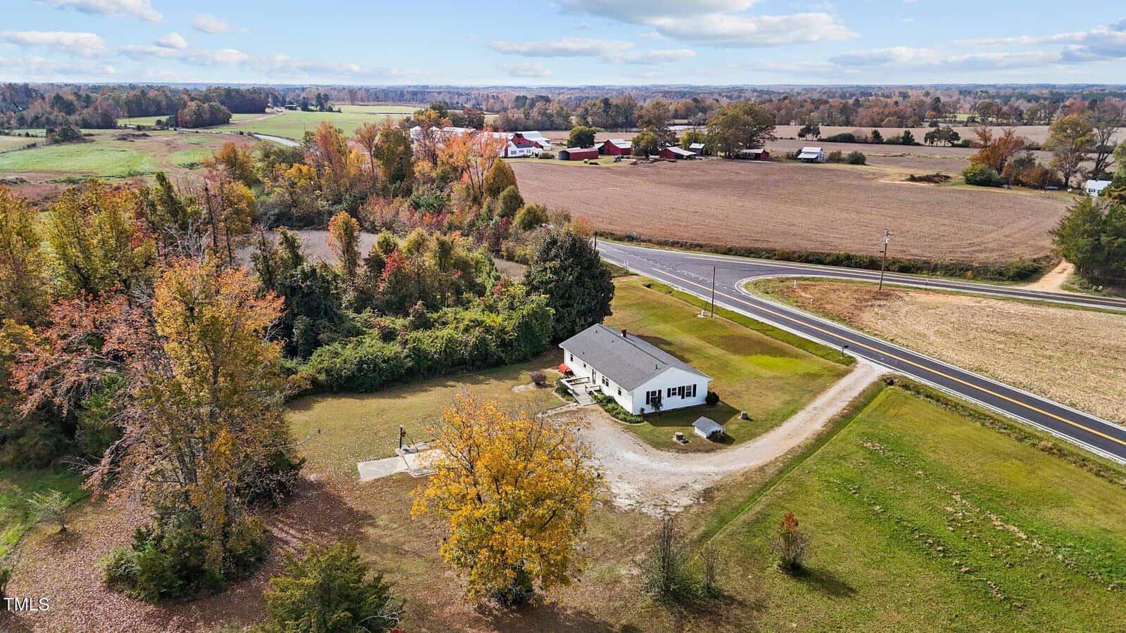 Aerial view of a rural landscape featuring a single-story white house with a gray roof surrounded by trees. A paved road curves near the property, and fields stretch into the distance under a partly cloudy sky.