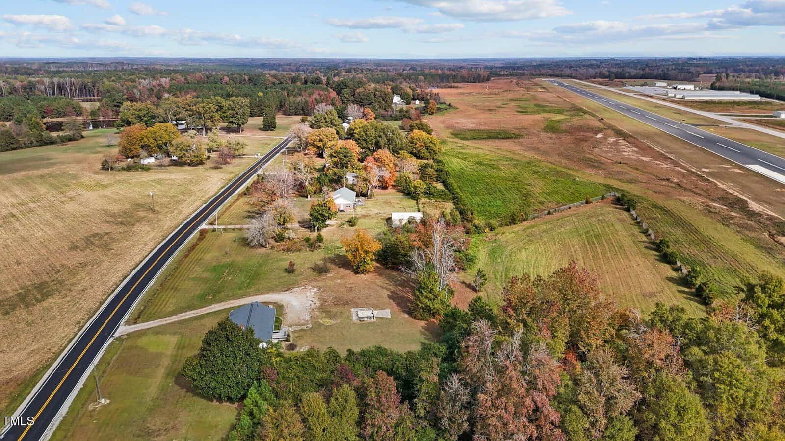 Aerial view of a rural landscape with a road running parallel to an airport runway. Surrounding areas include fields, scattered trees with autumn foliage, and several houses. The sky is partly cloudy.