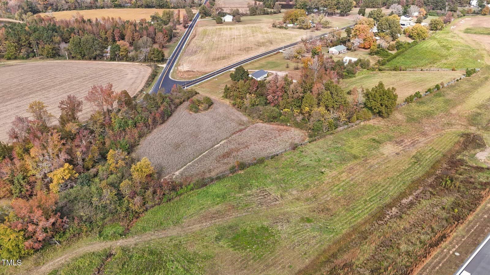 Aerial view of a rural landscape featuring a patchwork of fields and pastures. A paved road divides the area, flanked by trees in autumn colors. Residential houses are scattered among the fields.