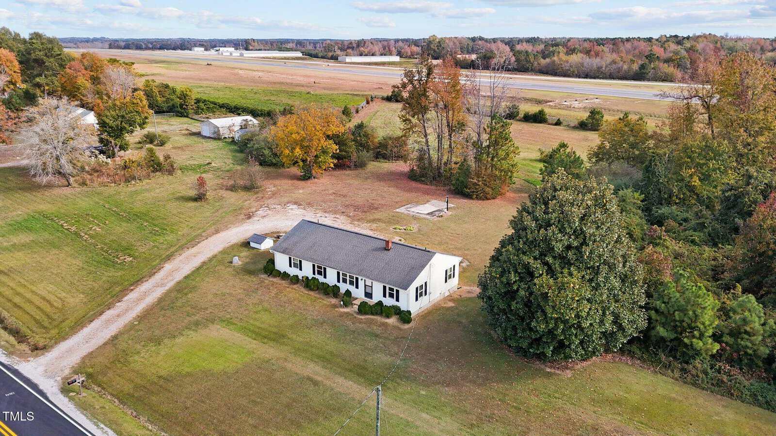 A small white house with a dark roof is surrounded by grass and trees in an open countryside setting. A dirt driveway curves from the road to the house. The background features fields, trees in autumn colors, and a clear sky.