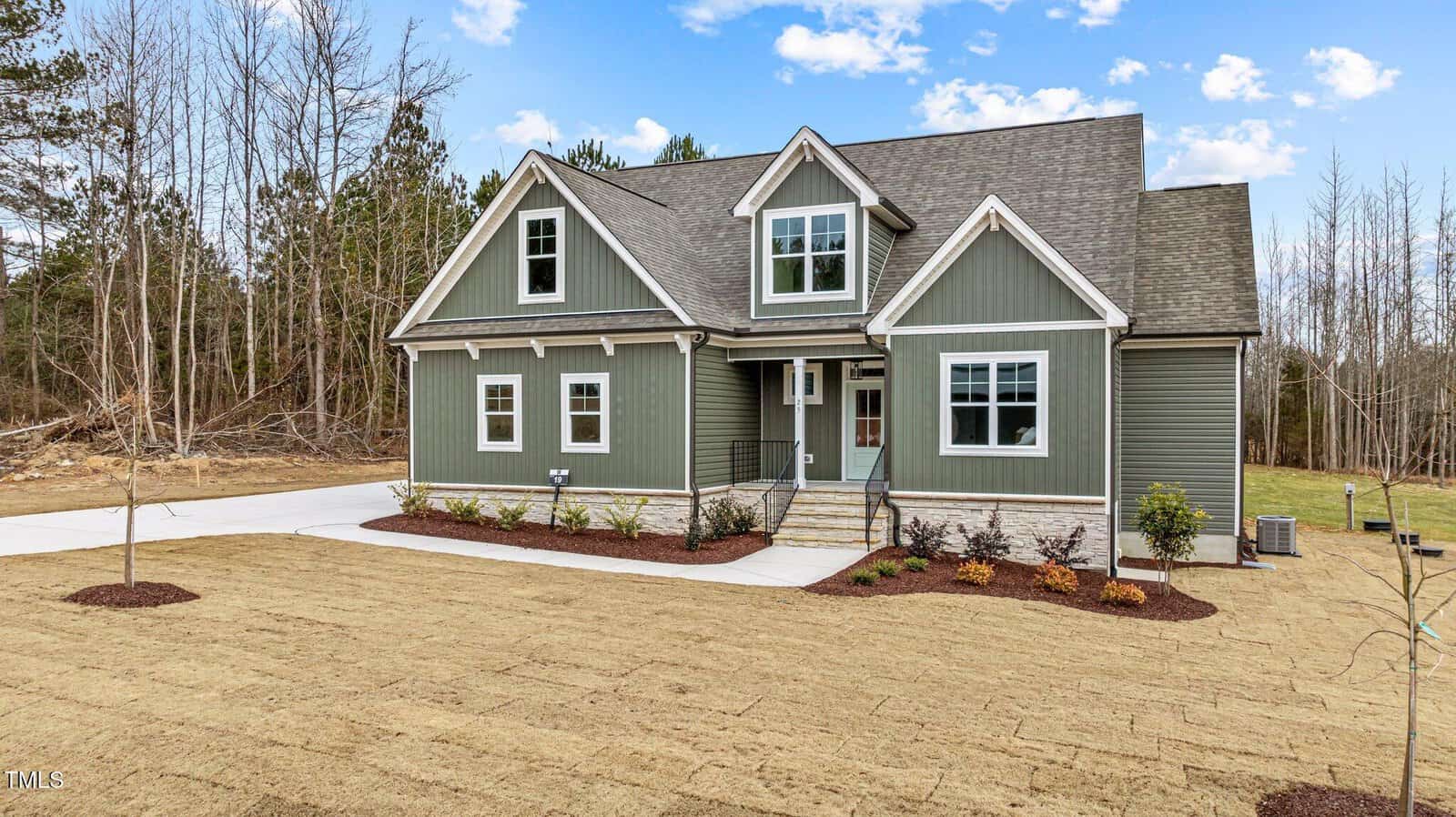 A green, two-story house with white trim and a front porch sits on a plot with freshly laid sod, young plants along the walkway, and a backdrop of leafless trees and evergreens under a partly cloudy sky.