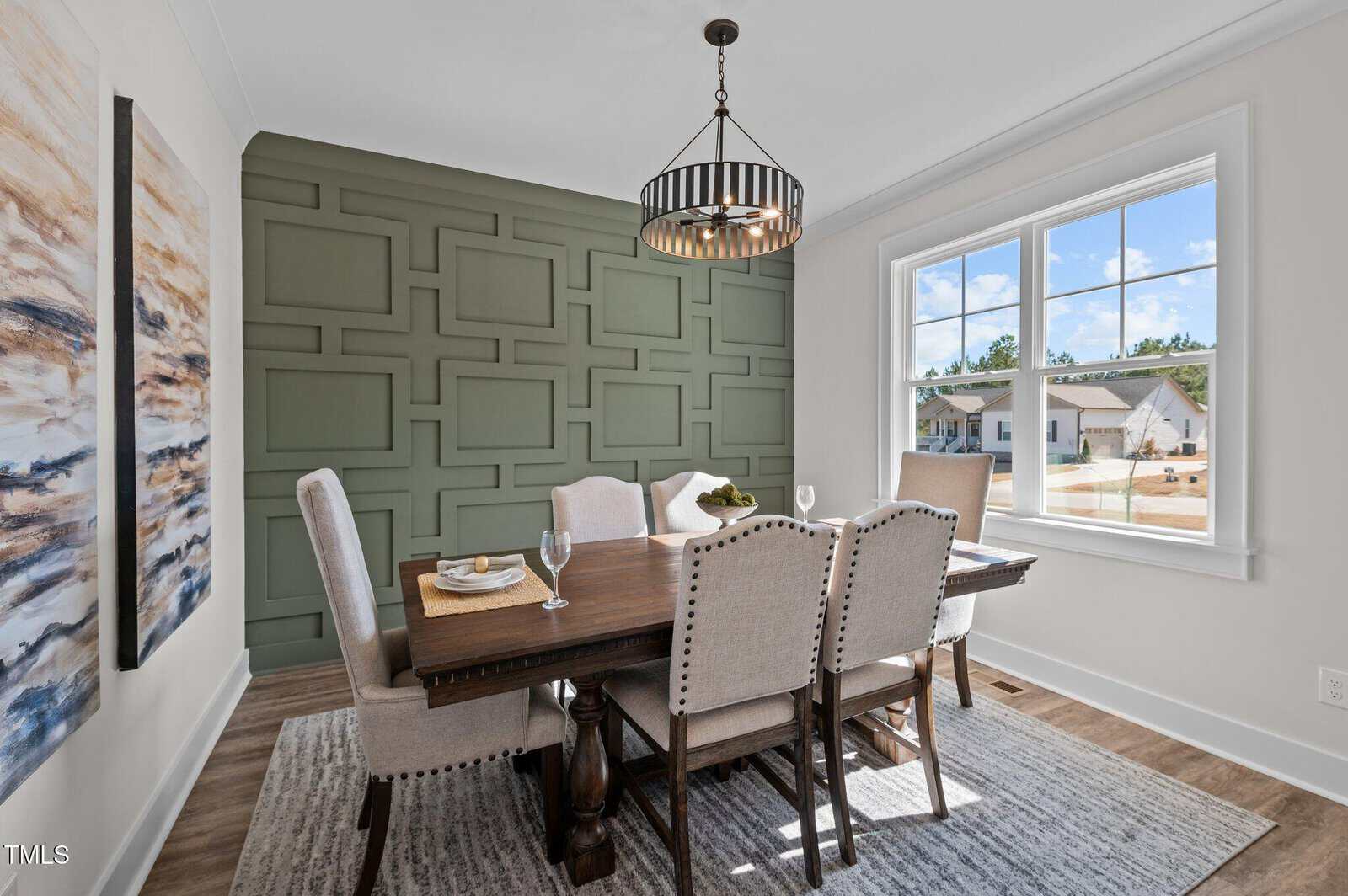 A dining room with a wooden table, six upholstered chairs, an abstract painting on the left wall, a geometric green accent wall, a modern light fixture, and a window showing a suburban neighborhood.