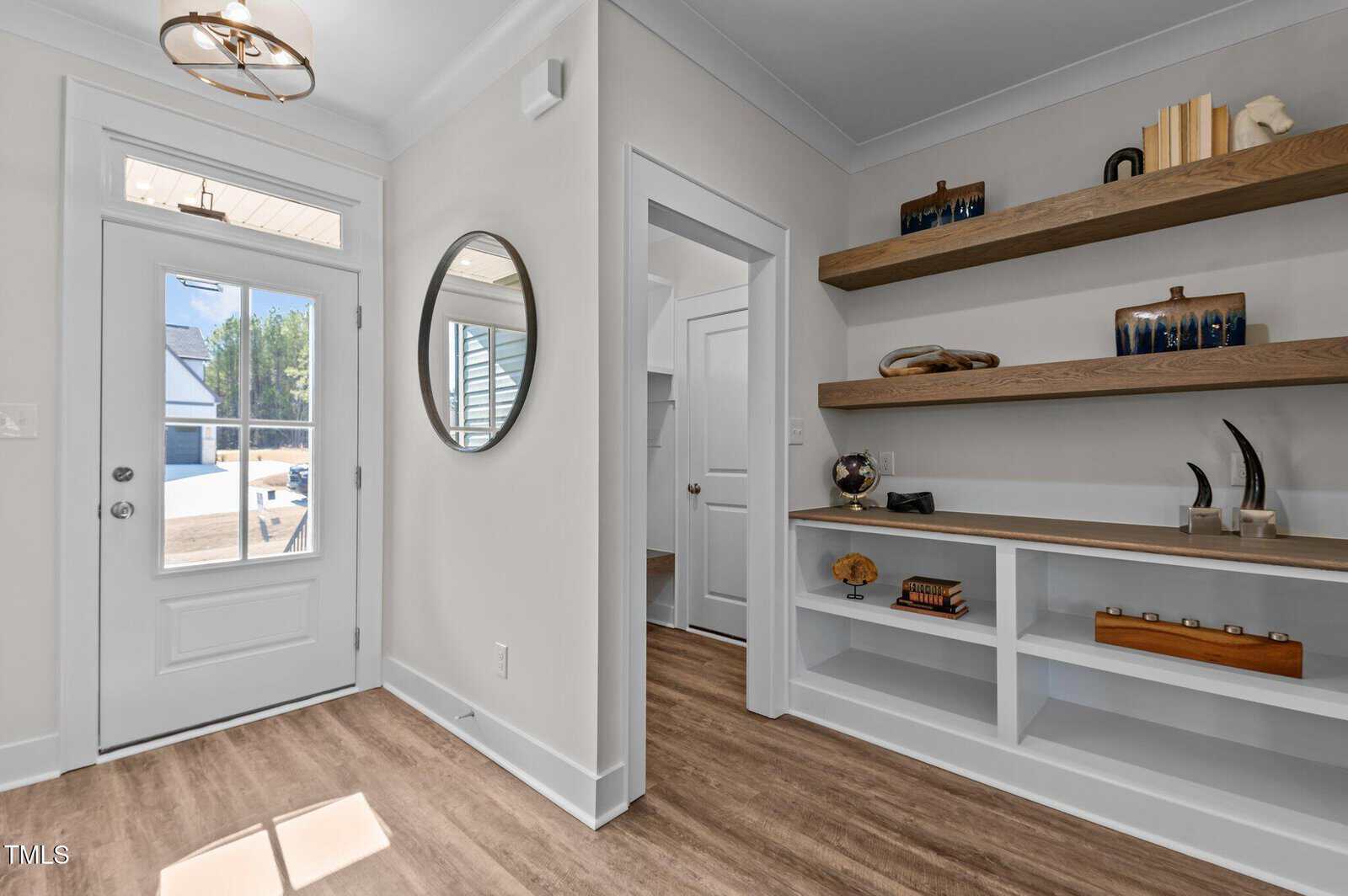 A modern entryway with light wood flooring, a white door with glass panes, a round mirror on the wall, and built-in shelves displaying books, decorative objects, and small sculptures.