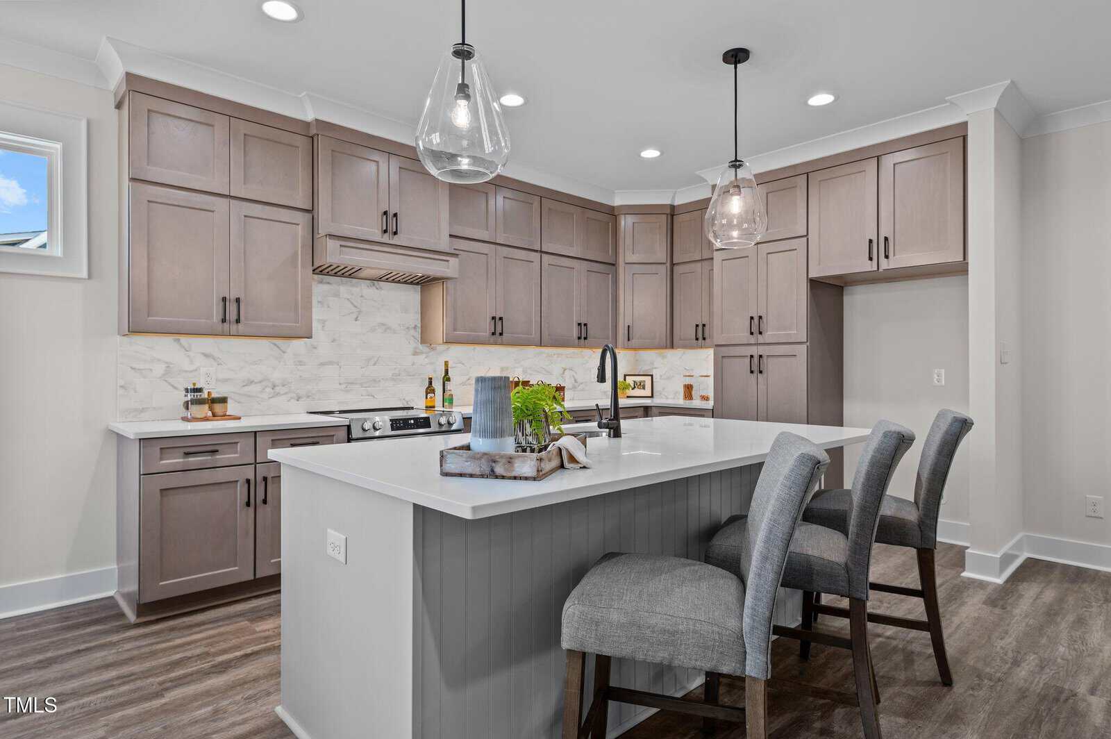 Modern kitchen with light brown cabinets, white marble backsplash, island with white countertop, two gray upholstered bar stools, stainless steel appliances, pendant lights, and wood flooring.