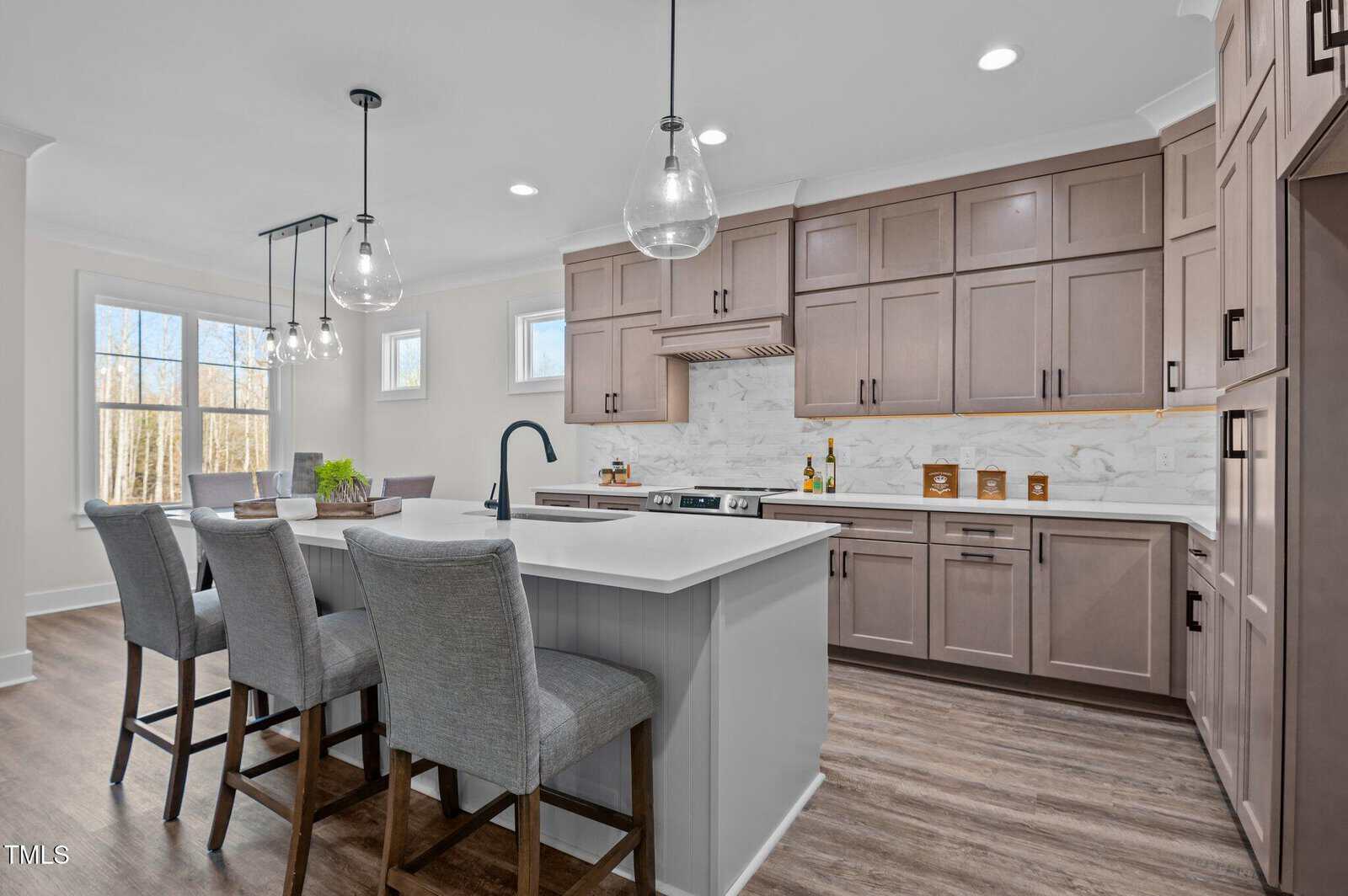 Modern kitchen with taupe cabinets, marble backsplash, kitchen island with gray upholstered stools, pendant lights, built-in appliances, and a dining area with a window showing trees outside.
