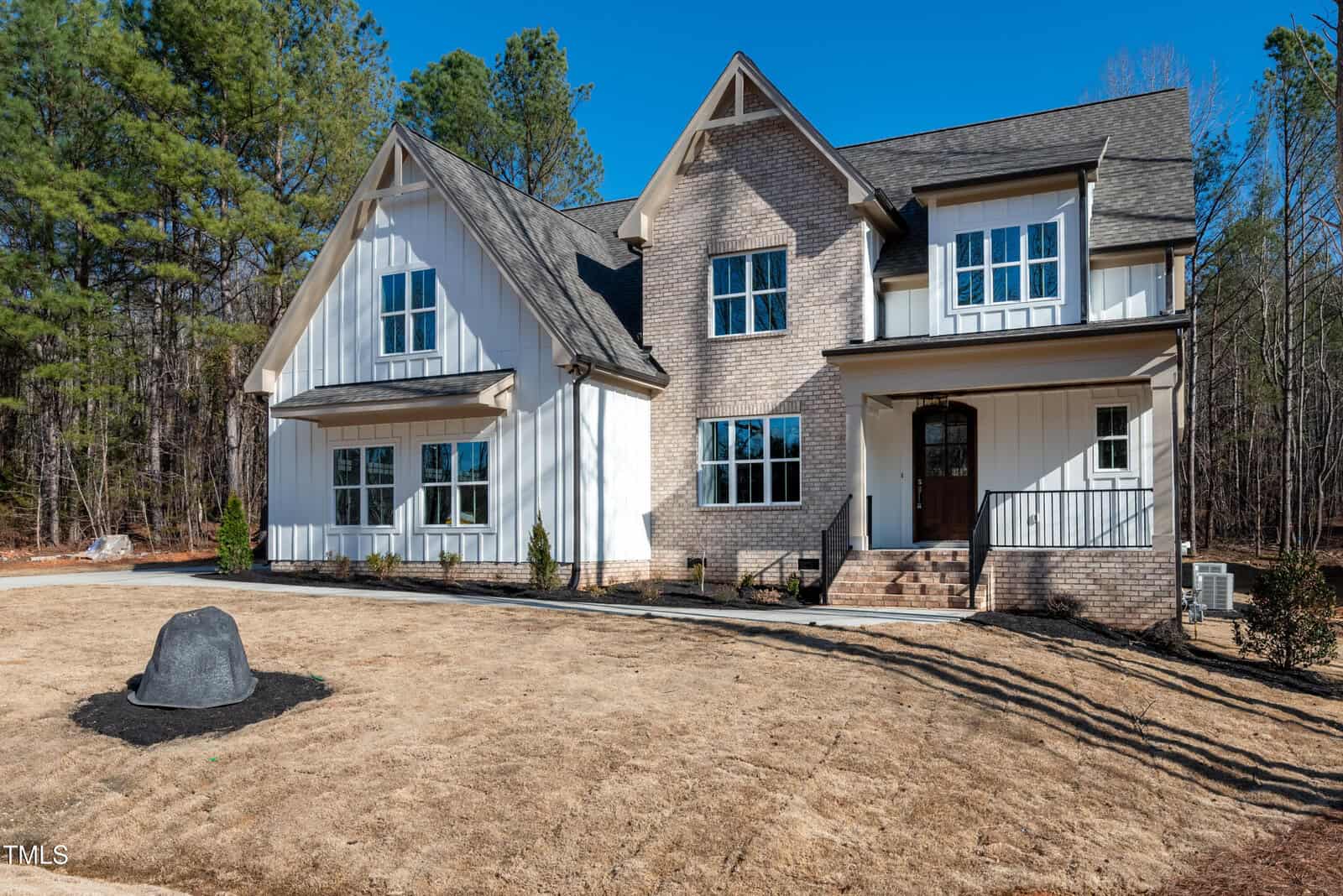 A modern two-story house with light brick and white siding, large windows, and a covered front porch. The yard is mostly brown grass with a single landscaped rock near the front, surrounded by trees.