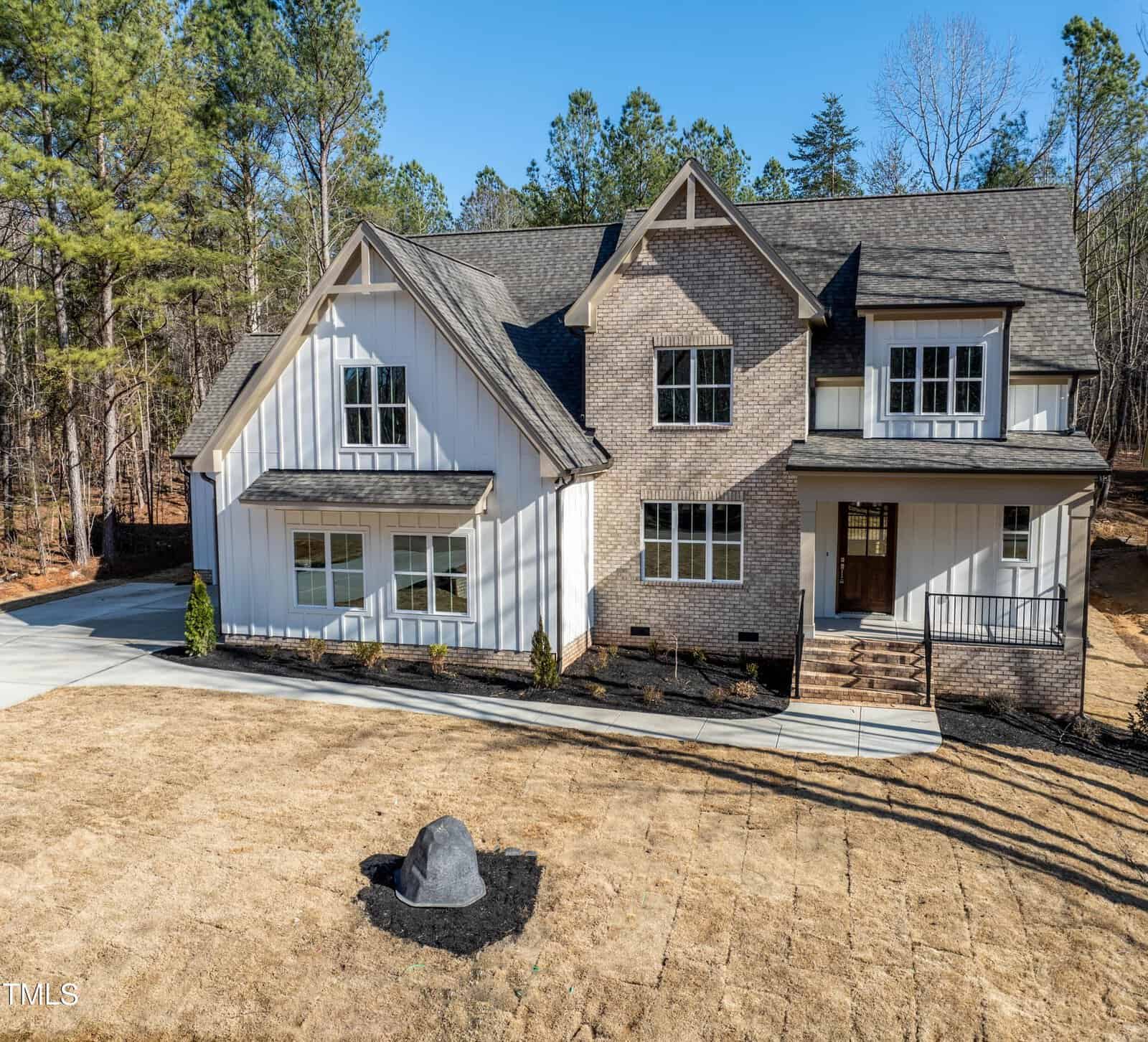 A large modern farmhouse-style home with a combination of white siding and brick exterior, black trim windows, and a covered front porch, surrounded by a dry lawn and trees in the background.