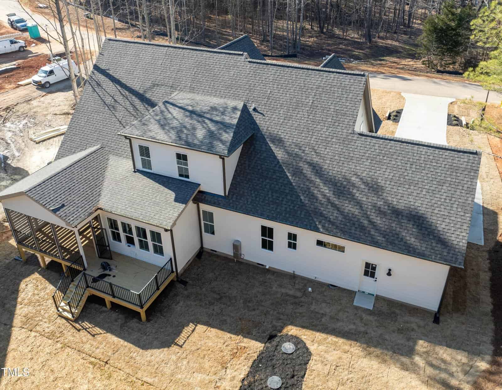 Aerial view of a large, modern house with dark gray shingles, white exterior walls, and a covered back porch with a wooden deck, surrounded by bare soil and some newly planted landscaping.