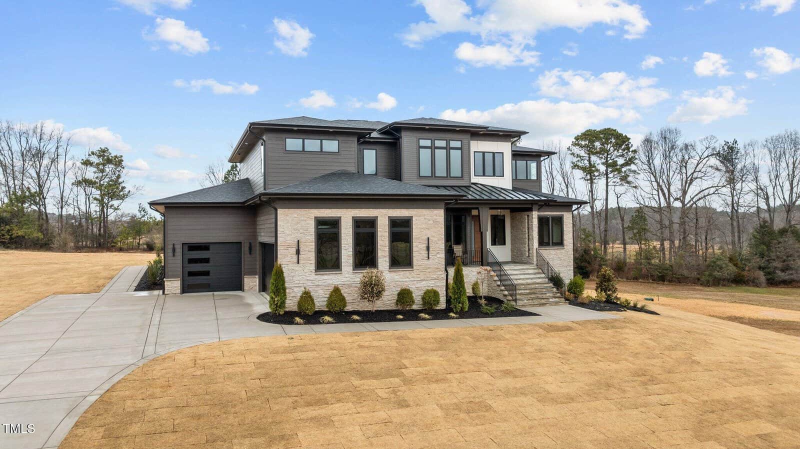 A modern two-story house with large windows, a stone and siding exterior, attached single-car garage, landscaped front yard, and a wide driveway, set against a backdrop of trees and an open field under a partly cloudy sky.