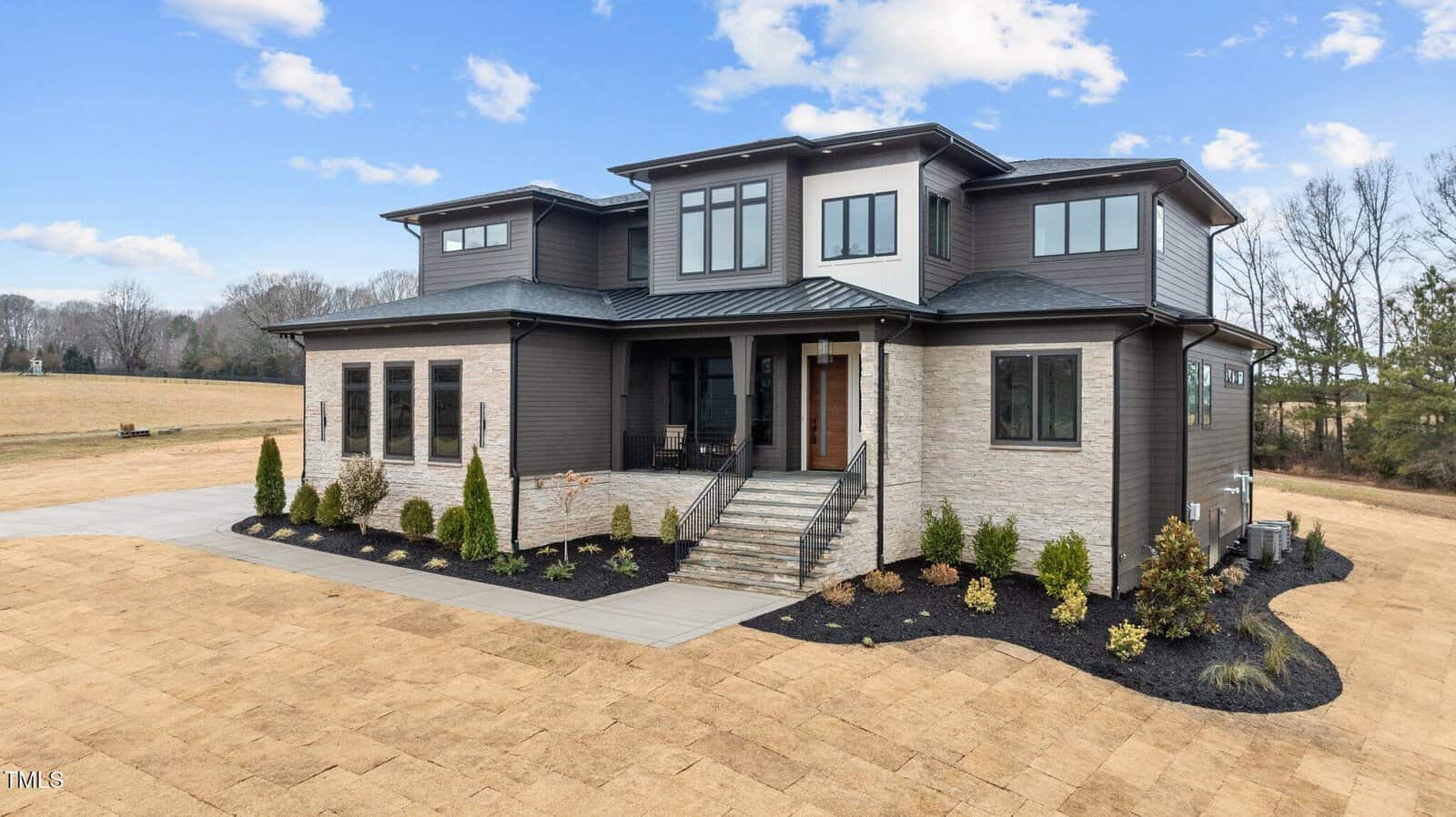 A modern two-story house with a mix of light stone and dark siding, large windows, a covered front porch with stairs, and landscaped shrubs, set on a spacious lot with a clear sky above.