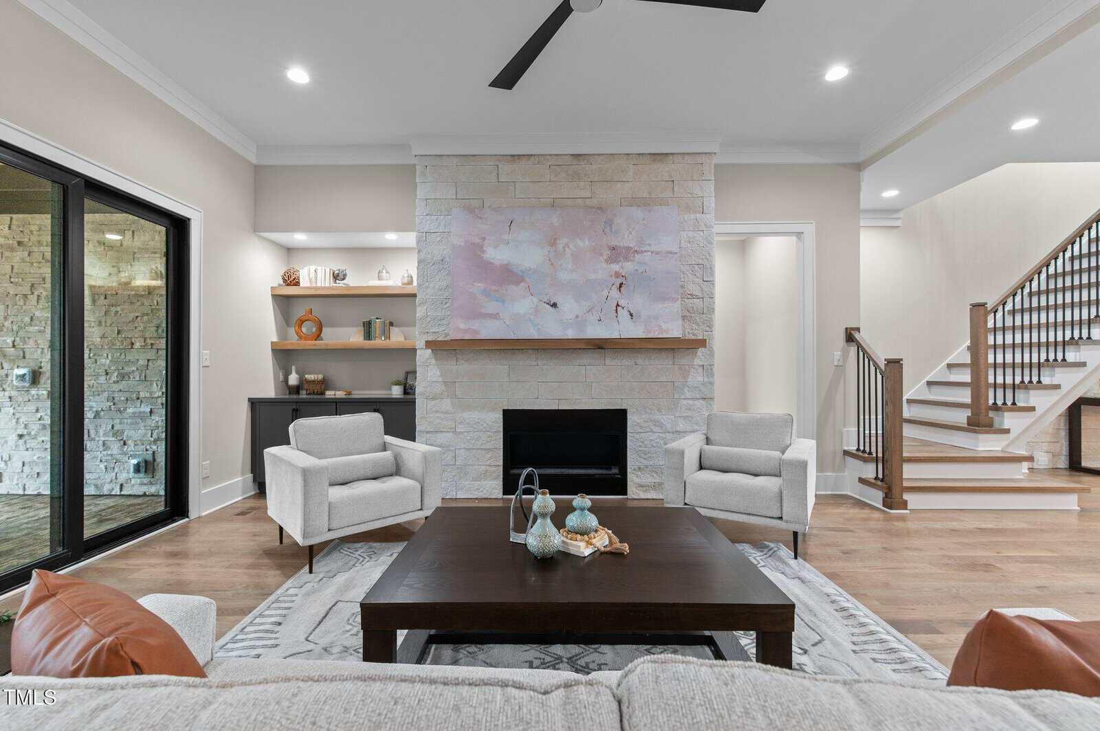 A modern living room with light walls, a stone fireplace with a large abstract painting above it, two armchairs, a dark wood coffee table, shelves to the left, and stairs to the right.