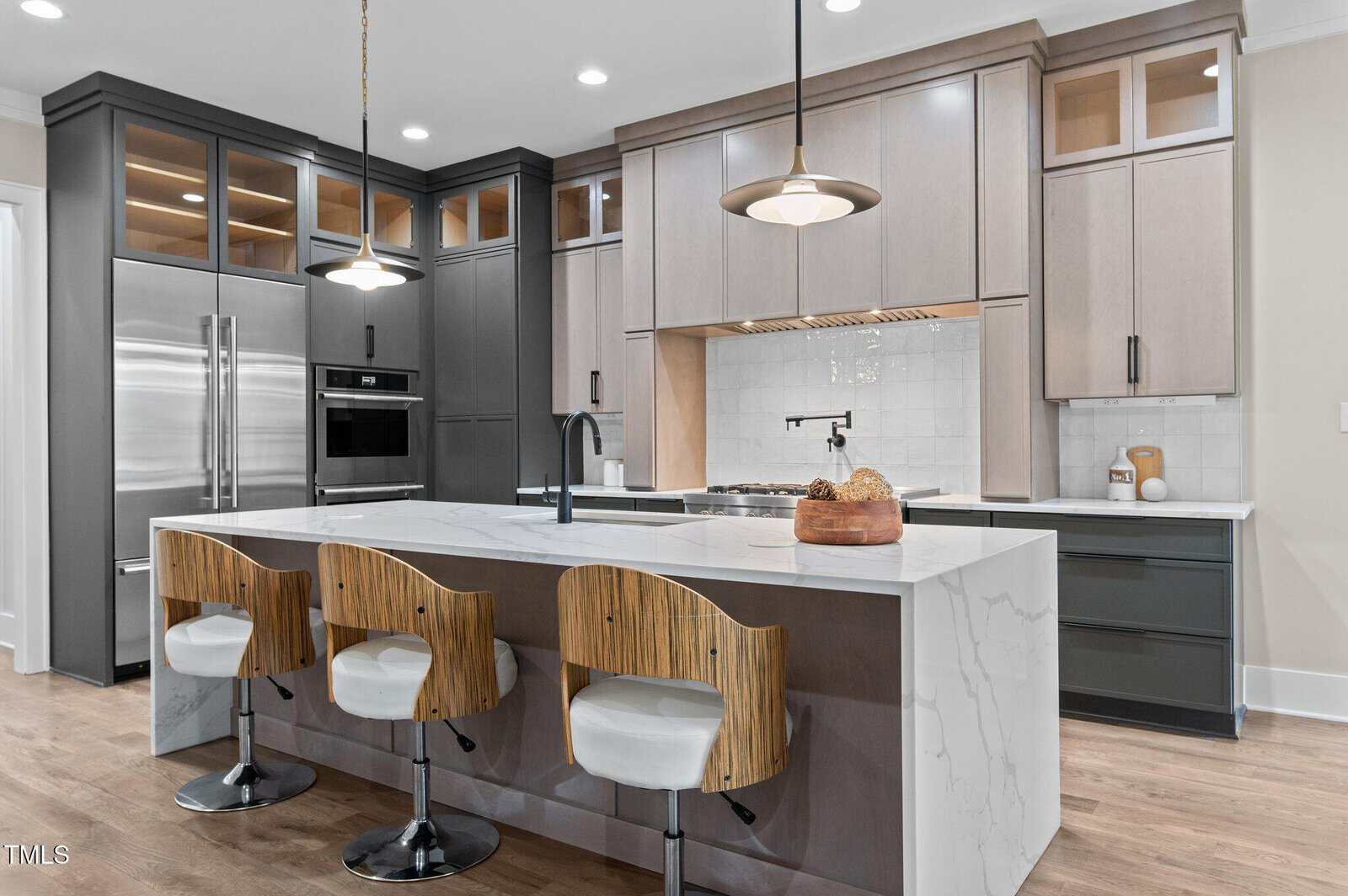 Modern kitchen with light wood flooring, a large white marble island with three wood and white cushioned bar stools, dark lower cabinets, light upper cabinets, double wall ovens, and pendant lighting.