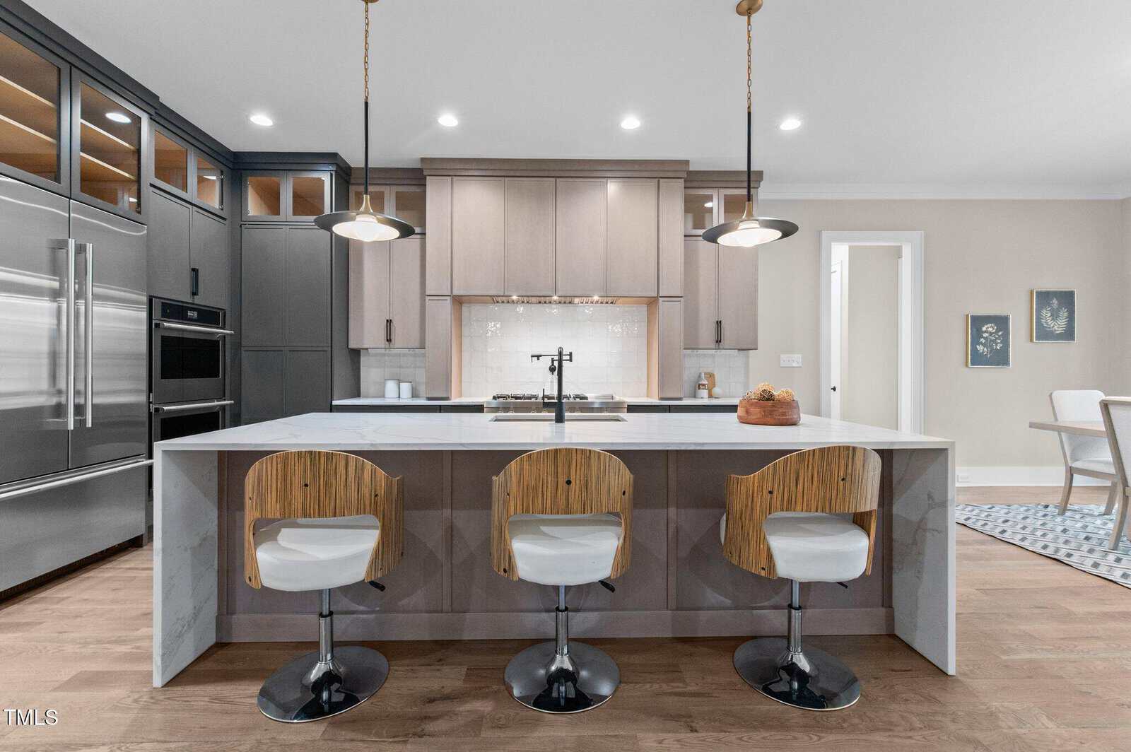 A modern kitchen features a large marble island with three wooden barstools, stainless steel appliances, pendant lights, and light wood cabinetry. A dining area with a table and chairs is visible in the background.