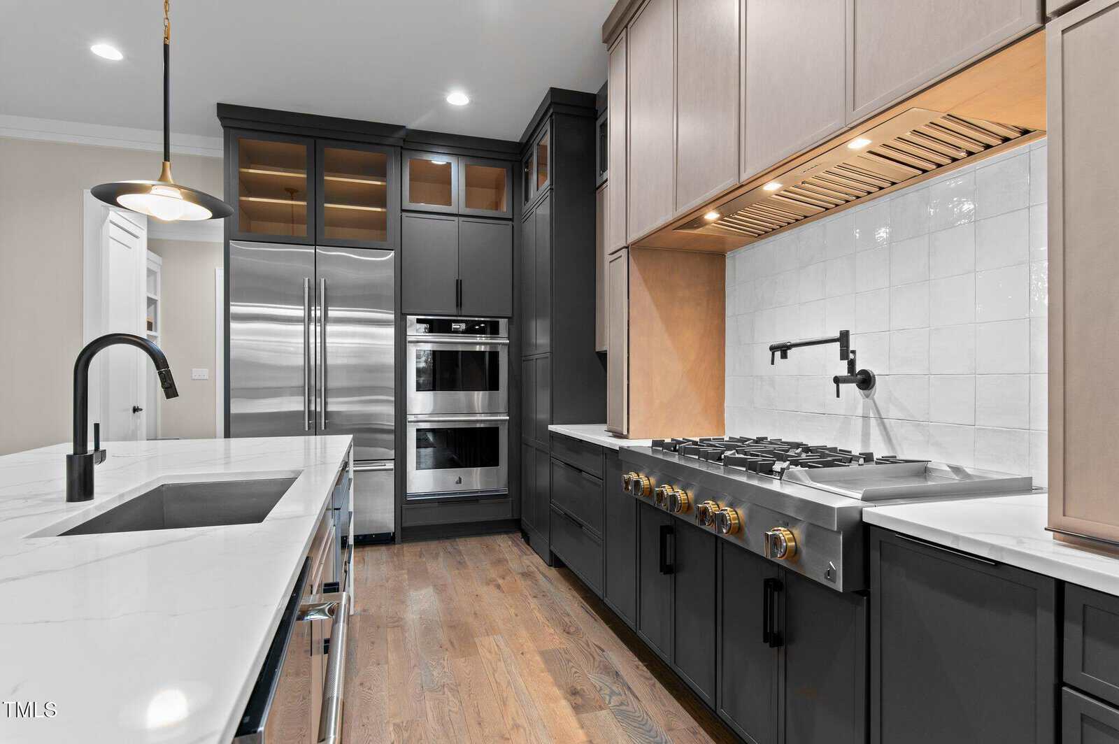 Modern kitchen with dark cabinets, stainless steel appliances, built-in oven, gas cooktop, white tile backsplash, marble countertops, undermount sink, and light wood flooring. Overhead lighting and open shelves are visible.