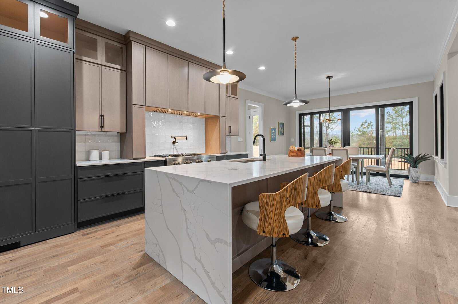 Modern kitchen with a large marble island, three wooden barstools, pendant lights, light wood flooring, and black and wood cabinetry. A dining area with a table and chairs is visible near large windows in the background.