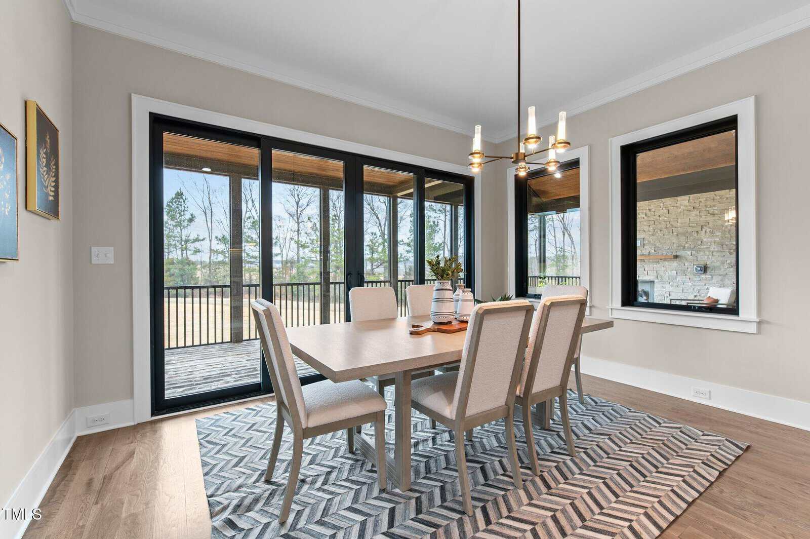 A modern dining room with a light wood table and six beige upholstered chairs on a geometric rug. Large glass doors and windows offer a view of a patio and trees outside. A chandelier hangs above the table.
