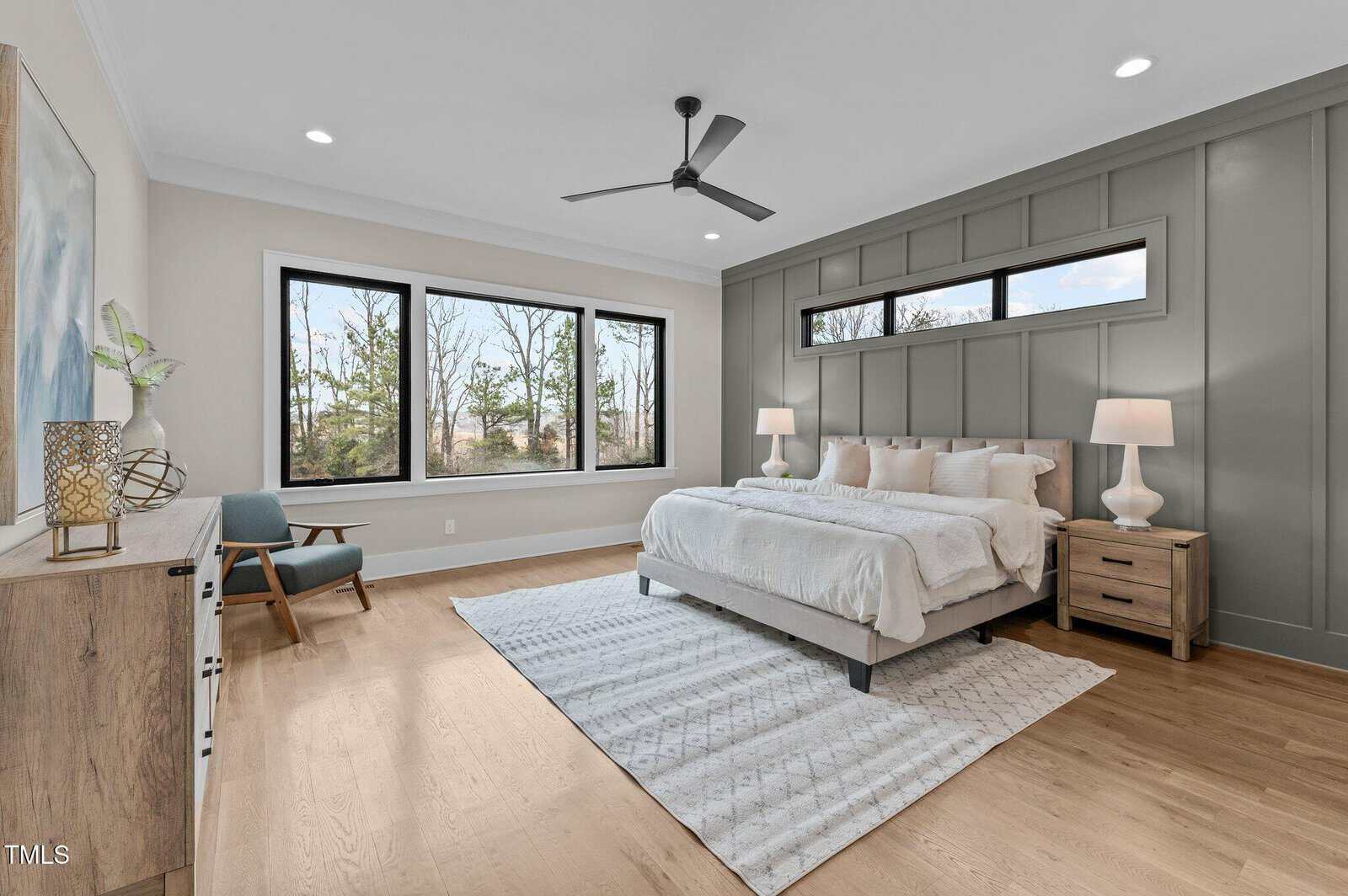Modern bedroom with large windows, a bed with white bedding, two bedside tables with lamps, a wooden dresser, and a gray accent wall. Light wood flooring and a patterned white rug complete the space.