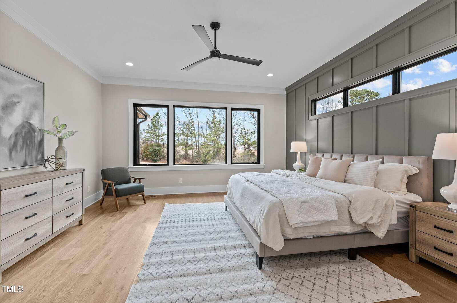 Modern bedroom with large windows, gray accent wall, king-sized bed with white bedding, two nightstands with lamps, dresser, armchair, ceiling fan, and light wood flooring with a patterned rug.