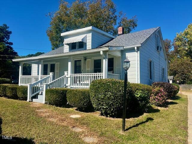 A small, white, single-story house on 203 Peace Street features a covered front porch with steps leading up. Surrounded by neatly trimmed bushes and a light post in the Oxford front yard, the home sits under a clear blue sky with trees in the background.