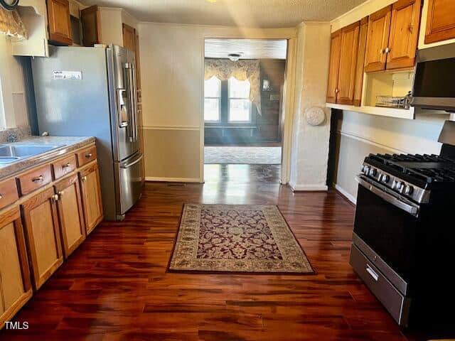 A kitchen with wooden cabinets, stainless steel refrigerator, and a gas stove. A patterned rug covers the wooden floor. The view extends into an adjacent room with large windows and a brown wall. A smoke detector is visible on the ceiling.