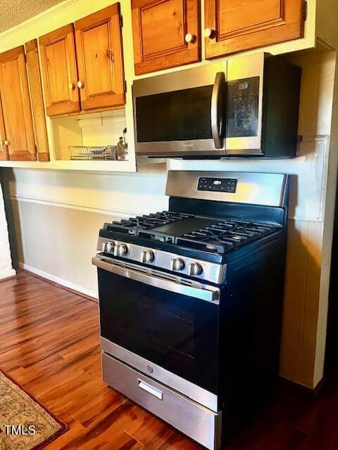 A kitchen features stainless steel appliances, including a gas stove with an overhead microwave. Wooden cabinets are above, and the floor is laminated with a wood finish. A small rack with dishes is on the left countertop.