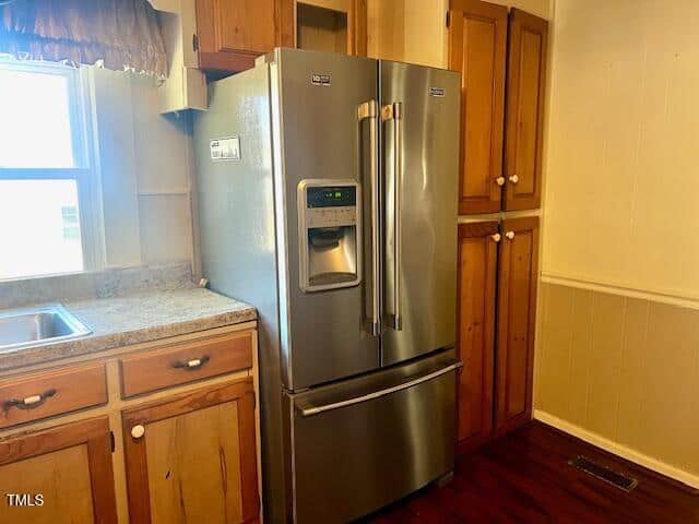 A stainless steel French door refrigerator with an ice and water dispenser is placed next to wooden kitchen cabinets. The kitchen countertop is granite, and a single sink is visible beside the fridge. Sunlight filters through a window above the sink.